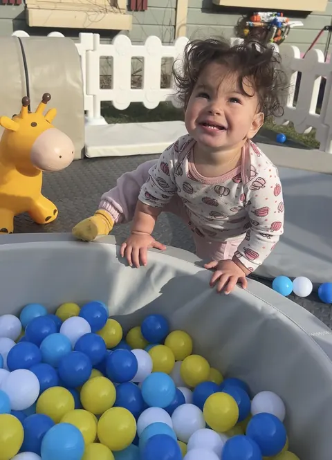 A smiling toddler playing in a Little Giggles ball pit full of blue, yellow and white balls